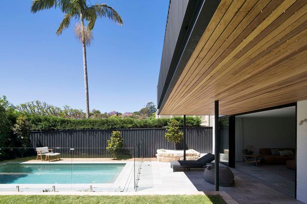 A contemporary residential poolside exterior with a long cantilevered or overhanging roof clad in warm horizontal timber boards. The image shows a turquoise swimming pool, pale paved pool surround, dark boundary fence, landscaped garden, palm tree, and a shaded open living area with dark-framed glazing and outdoor furniture.