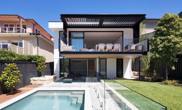 A contemporary multi-level residential rear facade with a covered upper balcony, black metal balustrades, extensive glazing, and a flat roof with projecting slatted sunshades. The ground level opens onto a landscaped lawn and paved terrace beside a rectangular swimming pool, with neighbouring homes visible on either side.