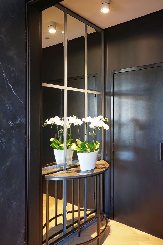 A refined dark-toned interior entryway featuring a black-framed mirrored glass partition, a narrow curved console table with a timber top and metal balustrade-style base, and white orchid arrangements. Black paneled walls, integrated lighting, and warm timber flooring create a dramatic yet elegant atmosphere.