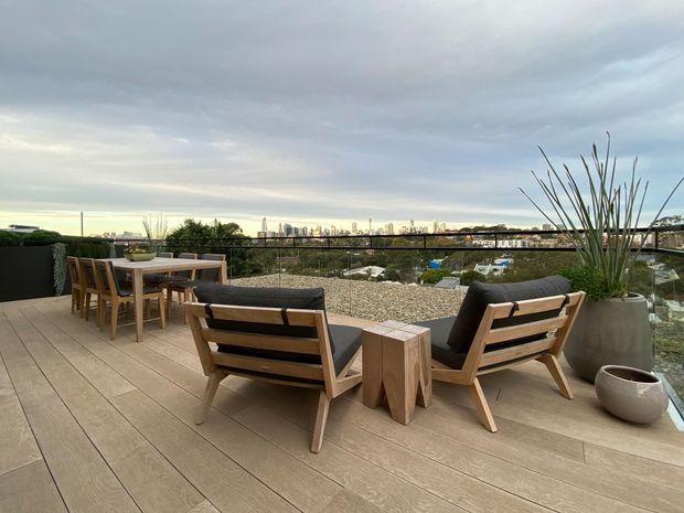 A contemporary rooftop terrace or balcony with a broad timber-look deck, outdoor dining table and chairs, and low wooden lounge seating arranged around a small side table. Large planters with architectural grasses frame the space, while a dark metal-and-glass balustrade overlooks a low-rise urban landscape and distant city skyline beneath an overcast sky.