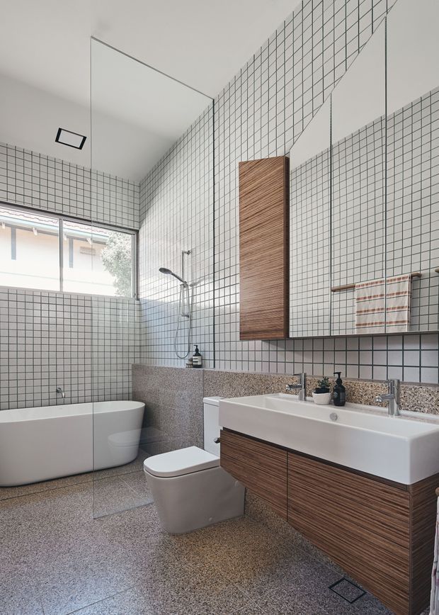 Contemporary bathroom with a freestanding white bathtub, wall-mounted toilet, broad white basin, frameless glass shower screen, and large mirrored cabinet. The walls are clad in small square white grid tiles with dark grout, while the floor, vanity benchtop, and bath surround use speckled terrazzo-like surfaces. Warm timber-grain vanity cabinetry and a matching tall recessed cabinet add contrast. The space is bright, clean, and highly geometric.