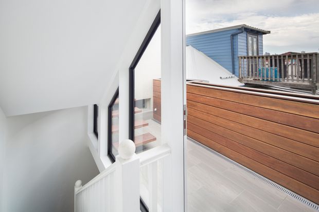 Bright contemporary stair landing beside full-height glazing, with a white painted stair balustrade and dark-framed windows. A timber-clad rooftop terrace or balcony is visible outside, along with neighbouring coastal-style buildings. The interior is clean and minimalist with strong natural light and contrasting warm timber outdoors.