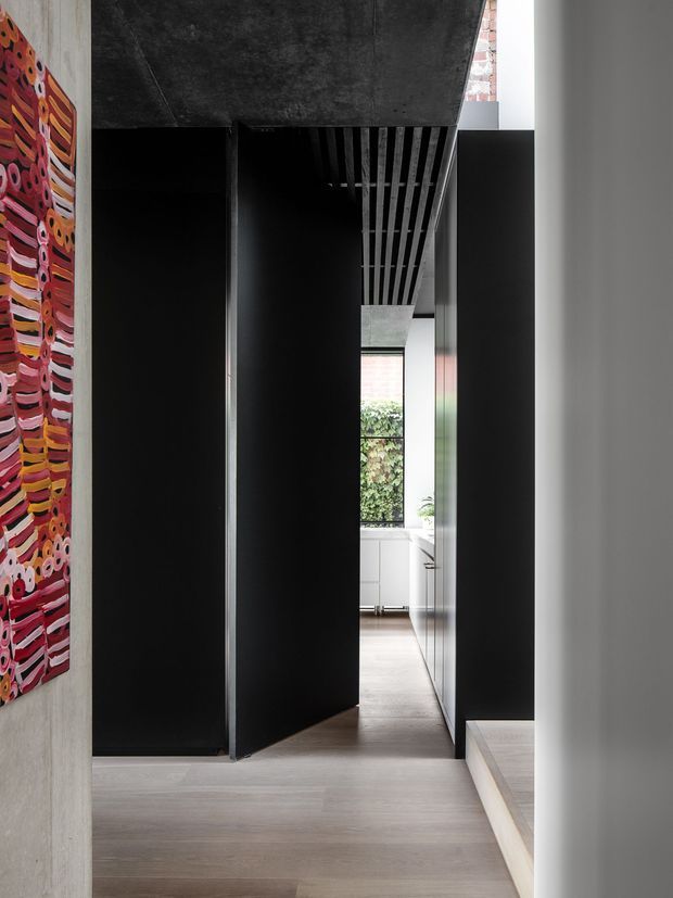 A narrow, dramatic interior passage with dark black built-in cabinetry or wall planes, pale timber flooring, and a charcoal textured ceiling. A large colourful abstract artwork hangs on the concrete-toned wall at left. Slatted ceiling detailing leads toward a brighter kitchen or living area with white cabinetry, glazing, and greenery beyond. The composition has strong linear perspective and high contrast between the dark corridor and bright destination space.