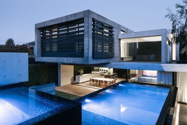 Modern multi-level concrete residence photographed at dusk, organized around a bright blue swimming pool. The house features strong cantilevered volumes, exposed concrete framing, extensive glazing, dark horizontal louvres, and open-plan living and dining areas visible beneath the upper level. The composition emphasizes indoor-outdoor connection, geometric form, and integrated pool terraces.