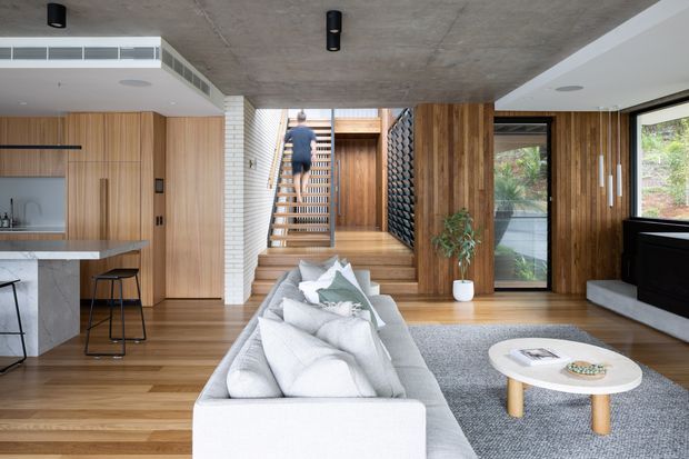 A contemporary open-plan living area with a large pale sectional sofa in the foreground, a round light coffee table, timber flooring, and extensive timber wall panelling and cabinetry. A compact kitchen with a stone island and stools sits to the left, while a central timber staircase with open treads leads upward. The room features a raw concrete ceiling, dark cylindrical ceiling lights, a textured dark feature wall beside the stairs, and large glazed openings looking onto greenery.