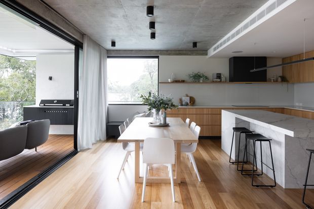 A bright, contemporary open-plan kitchen and dining area with warm timber flooring and cabinetry, a long marble-look island with black bar stools, and a light timber dining table with white chairs. Full-height glazing and a large sliding glass opening connect the interior to a balcony or outdoor area. The space features an exposed concrete ceiling, recessed linear air-conditioning grille, black accents, and minimalist pendant-style spot lighting.