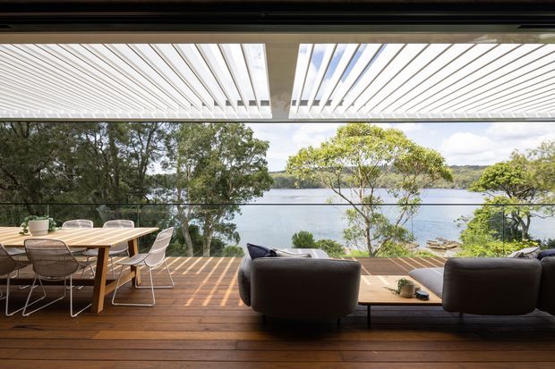 A waterfront outdoor living and dining area on a timber deck, viewed through an open glazed enclosure. The space features a timber dining table with wire-frame chairs, a dark upholstered sofa arrangement, frameless glass balustrading, and a white louvred pergola overhead. Trees and a calm body of water are visible beyond the deck.
