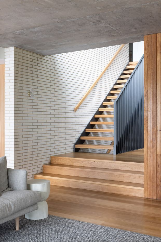 A bright modern stairway and landing viewed from a living area. The space features open timber stairs with a timber handrail, a dark vertically ribbed metal balustrade, white glazed brick walls, a raw concrete ceiling, warm timber flooring and wall panelling, and a grey upholstered armchair with a small round side table in the foreground.