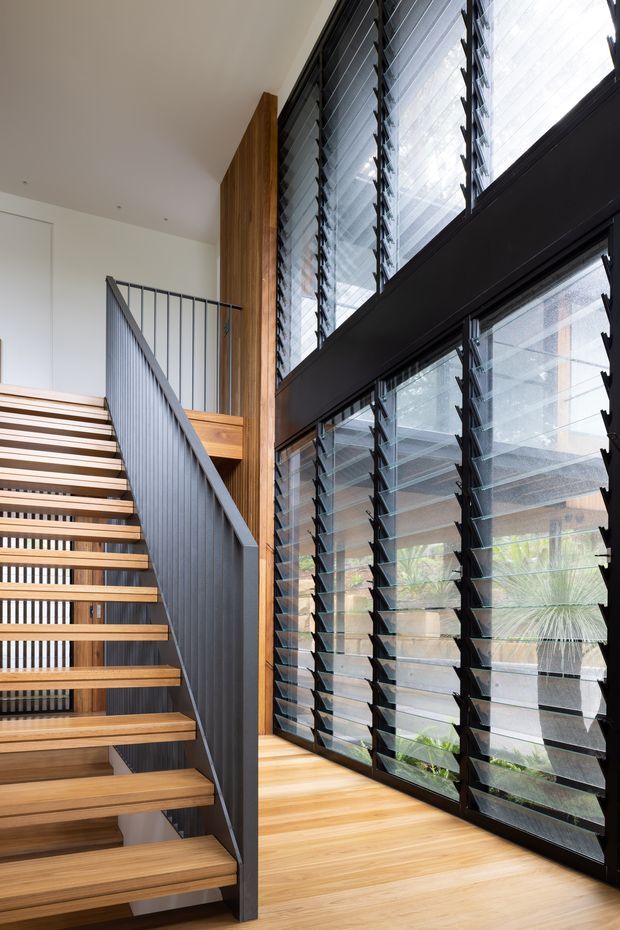 Bright contemporary interior stair hall featuring an open-riser timber staircase with dark metal balustrade, a tall exposed timber wall element, and expansive black-framed louvred glazing. The warm timber floor and stairs contrast with white walls and dark window framing, creating an airy, vertical circulation space with strong natural light.