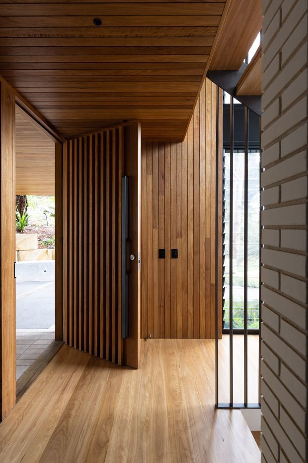 A contemporary residential entry hall with warm timber lining across the ceiling, walls, slatted screen door, and timber flooring. A tall dark-framed glazed opening brings in daylight beside a white exposed-brick wall, while the partially open front door connects to an exterior path and landscaping.