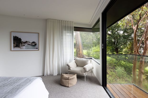 A bright, serene bedroom with a partially visible bed, soft grey carpet, white walls, sheer floor-to-ceiling curtains, and a compact upholstered lounge chair beside a corner of expansive black-framed glazing. The glazing opens onto a timber deck overlooking dense greenery, creating a strong indoor–outdoor connection. The room is simply styled with neutral textiles, a small woven ottoman, and understated framed artwork.