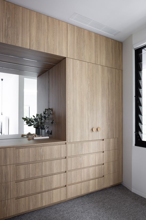 A built-in floor-to-ceiling timber-look storage and wardrobe wall dominates the room, with numerous drawers, cupboards, and a recessed open shelf containing a mirror and potted plant. The joinery has a light natural wood-grain finish with simple round knobs. A black-framed louvred window is visible on the right, and grey carpet covers the floor. The space is bright, clean, and highly organised.