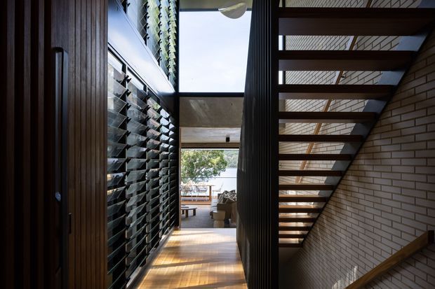 A narrow, sunlit interior circulation space with a timber floor and dark vertical timber wall panelling. The left side features a large louvred glazing or ventilation screen and tall built-in timber joinery, while an open-riser timber stair rises along a pale brick wall on the right. Daylight enters through high-level glazing and a skylight-like opening, with a glimpse of a living area beyond.