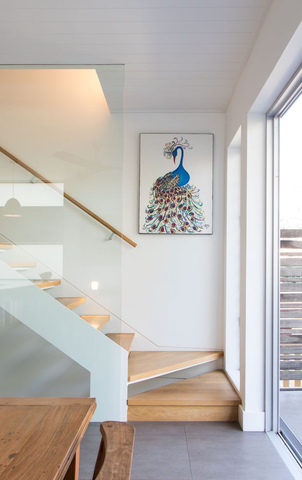 Bright contemporary stair hall and dining area with a sculptural timber staircase, clear glass balustrade, white-painted walls and ceiling, large glazed opening, and warm timber treads and handrail. A colourful peacock artwork provides a focal point on the wall, while a timber dining table is partially visible in the foreground.