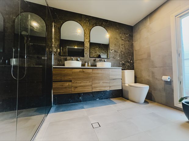 Contemporary bathroom with a frameless glass walk-in shower, double vanity with warm wood cabinetry and white basins, wall-mounted mirrors with arched tops, a white toilet, and large-format light stone-look floor and wall tiles. A dark textured stone feature wall creates contrast behind the vanity and shower.