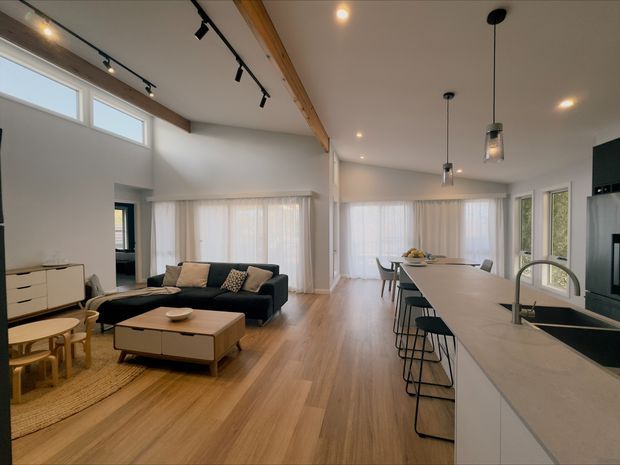 Open-plan contemporary kitchen, dining, and living area with warm timber flooring, white walls, extensive sheer-curtained glazing, and a vaulted ceiling with exposed timber beam. A long pale stone kitchen island with integrated sink and bar seating dominates the foreground, while a dark sectional sofa and timber coffee table define the living zone. Track lighting and pendant lights provide modern illumination.