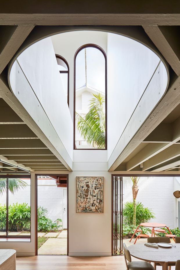 Bright interior looking toward a landscaped courtyard, with a dramatic double-height void and large arched clerestory window. Exposed painted timber beams frame the ceiling, while full-height glazing and glazed doors connect the room to the garden. A dining area with a round table occupies the foreground, and an abstract artwork hangs on the central wall. The palette is light and warm with timber flooring, white walls, dark timber window frames, and abundant greenery visible outside.