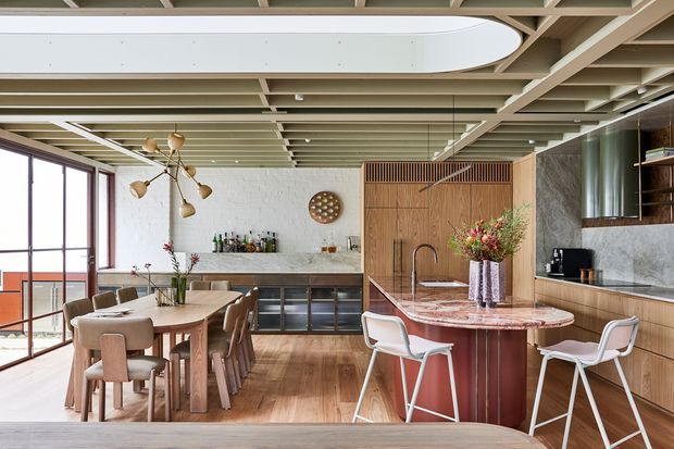 A spacious open-plan kitchen and dining area with a large timber dining table, upholstered dining chairs, timber kitchen cabinetry, a stone backsplash and island, and a sculptural marble-topped round island or bar. Full-height glazing opens to a balcony, while an exposed decorative timber ceiling grid with a large oval skylight-like feature creates a strong architectural focal point. The palette is warm timber, cream, muted green, and pink-red stone, with contemporary mid-century-inspired furnishings and brass-toned lighting.