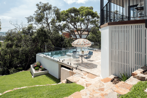 A contemporary multi-level residential outdoor entertaining area on a sloped site, with a stone-paved terrace, glass pool or deck balustrade, outdoor dining furniture, a patterned umbrella, and a modern white-clad building with vertical slatted screening and black-framed glazing. Landscaped lawn, mature trees, and distant urban views surround the space.