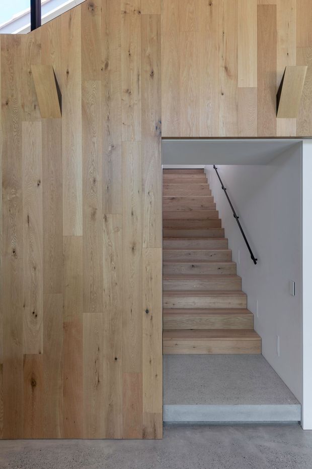 A minimalist interior stairway viewed from a concrete-floored lower level. Broad vertical oak timber wall cladding forms a warm feature surface around an opening to a straight flight of timber stairs. The stairs are bordered by smooth light-grey walls and a slim black handrail, with a concrete landing and a narrow high window providing natural light.