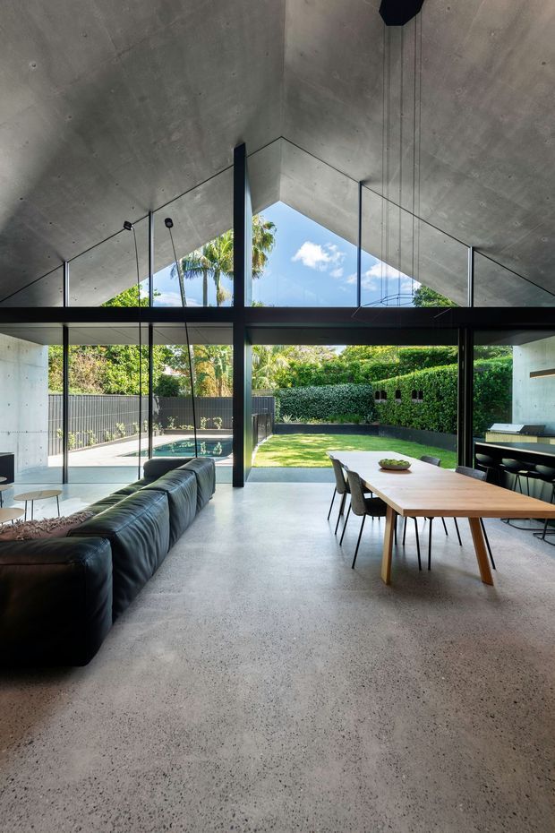 A dramatic open-plan modern living and dining space with a dark leather sectional, timber dining table, and integrated kitchen counter. Full-height glazing opens directly to a landscaped backyard and pool, while a sculptural exposed concrete vaulted ceiling and dark structural framing define the architecture. Pendant lights hang over the dining area, and the space has a strong indoor-outdoor connection with abundant natural light.