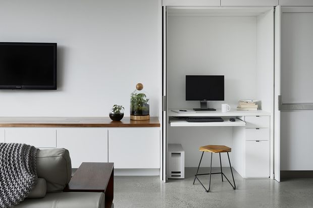 A clean, minimalist open-plan living area with a built-in white media unit and low timber-topped cabinetry. A wall-mounted television and decorative objects sit above the cabinetry, while a concealed home office nook contains a desk, monitor, keyboard, shelving, and a small timber stool. A grey sofa with a knitted throw is partially visible in the foreground, and polished grey concrete flooring extends across the space.