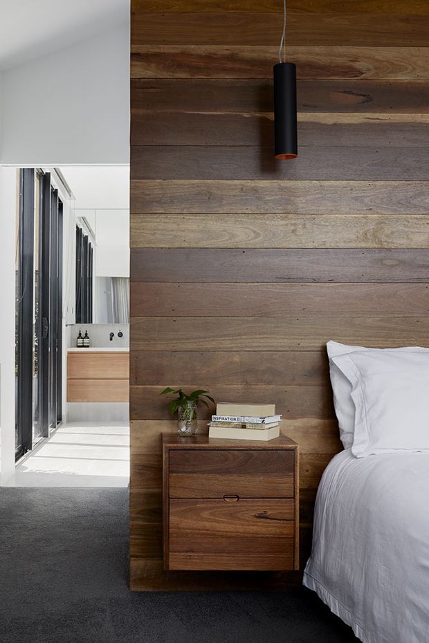 A contemporary bedroom featuring a full-height horizontal timber feature wall, a dark timber bedside table, white bedding, and a slim black pendant light. The bed sits beside a bright glazed corridor or bathroom area with dark-framed windows and pale built-in cabinetry. The room has a restrained, natural material palette and strong daylight.