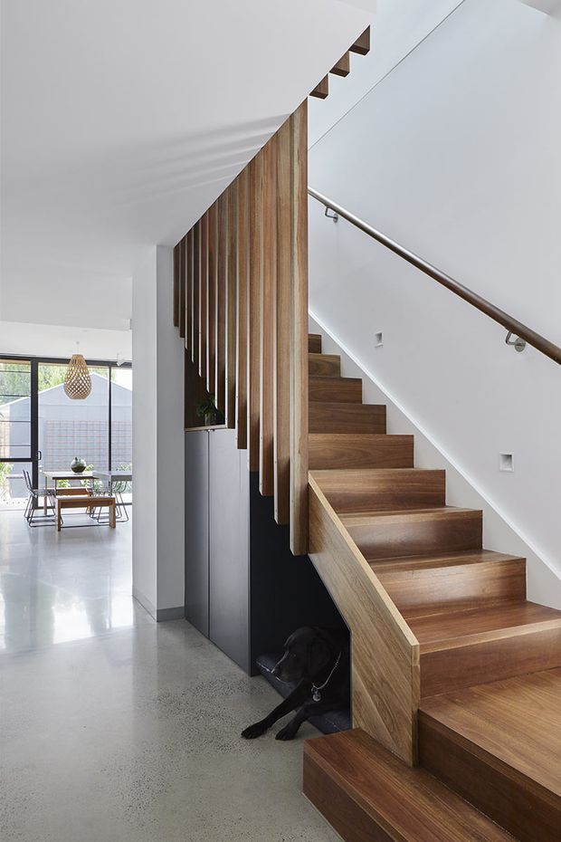 Interior view of a contemporary residential stairway with warm timber treads and a matching timber side stringer, set against crisp white walls and a dark under-stair joinery element. Vertical timber slats form a partial screen beside the stairs, while a slim timber handrail with metal brackets follows the white balustrade. A polished grey concrete floor extends toward a bright dining area with large glazing and a pendant light in the background.