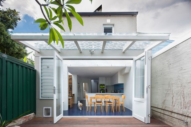 Bright view from an outdoor deck through wide-open glazed doors into a compact contemporary dining and kitchen area. A white-painted brick facade and covered pergola with patterned translucent panels frame the opening, while a timber dining table and chairs sit on a dark blue floor inside. Green fencing and surrounding planting border the narrow outdoor approach.