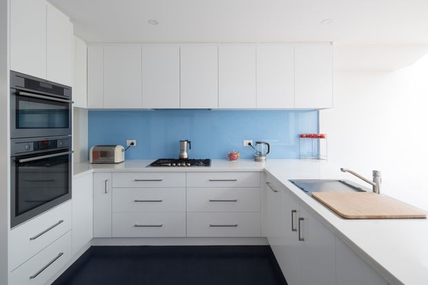 A bright, compact contemporary kitchen with a white U-shaped layout, extensive handleless and bar-handled cabinetry, integrated double wall ovens, a black cooktop, pale blue glass backsplash, and a white island or peninsula with an inset sink. The room has a clean, minimalist appearance with dark flooring providing contrast.