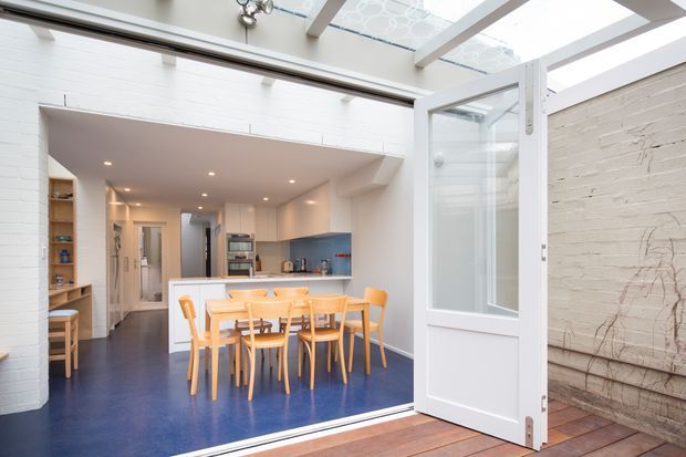 Bright open-plan kitchen and dining area viewed from a covered outdoor threshold through an open white-framed glazed door. The interior features white-painted brick walls, a vivid blue floor, pale timber dining furniture, white cabinetry, a blue backsplash, and exposed roof glazing that brings in abundant daylight. A rustic textured wall and timber decking are visible in the foreground and to the right.