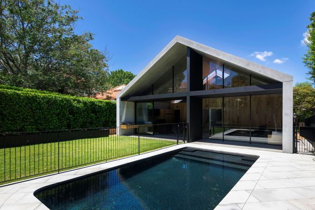 Modern single-storey residence with a dramatic asymmetrical pitched roof, dark framed floor-to-ceiling glazing, and a concrete roof and facade frame. The house opens onto a paved terrace and rectangular swimming pool, with a manicured lawn, hedge, and black metal pool fencing in the foreground.