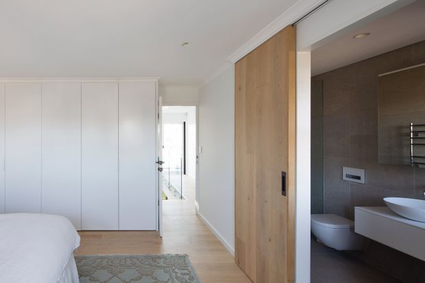 Bright contemporary bedroom with built-in white wardrobes, pale timber flooring, a large white bed, and a patterned rug. A wide timber sliding door opens directly to a modern ensuite bathroom with dark tiled walls, a wall-hung toilet, floating vanity, vessel basin, and heated towel rail.