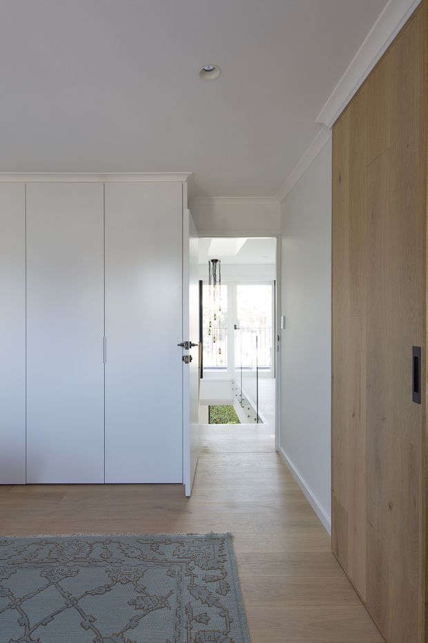 A bright, refined residential hallway or landing with pale timber flooring, white full-height built-in cabinetry on the left, and a large natural timber-clad wall or door on the right. The corridor leads toward a glazed stairwell or light-filled void with a contemporary pendant visible in the distance. White walls, simple cornices, recessed lighting, and a muted patterned rug create a clean, understated interior.