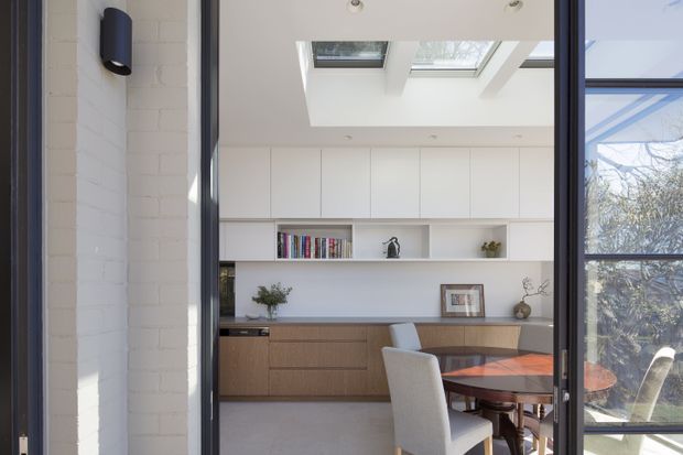 Bright contemporary dining and kitchen area viewed through a dark-framed glazed opening. The space features white built-in cabinetry with open shelving, warm timber-toned lower cabinetry, a dark benchtop, a timber dining table with upholstered chairs, and a large roof skylight bringing in natural light. White painted brickwork and extensive glazing are visible around the opening.