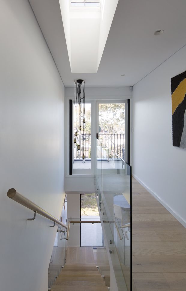 Bright contemporary stairway and landing viewed from above, featuring pale timber stair treads and flooring, white walls and ceiling, a large linear skylight, glass balustrades with dark metal framing, and a simple timber wall handrail. A tall narrow pendant light hangs through the stairwell, while glazed doors at the landing open toward an exterior balcony. Abstract wall art is partially visible on the right.