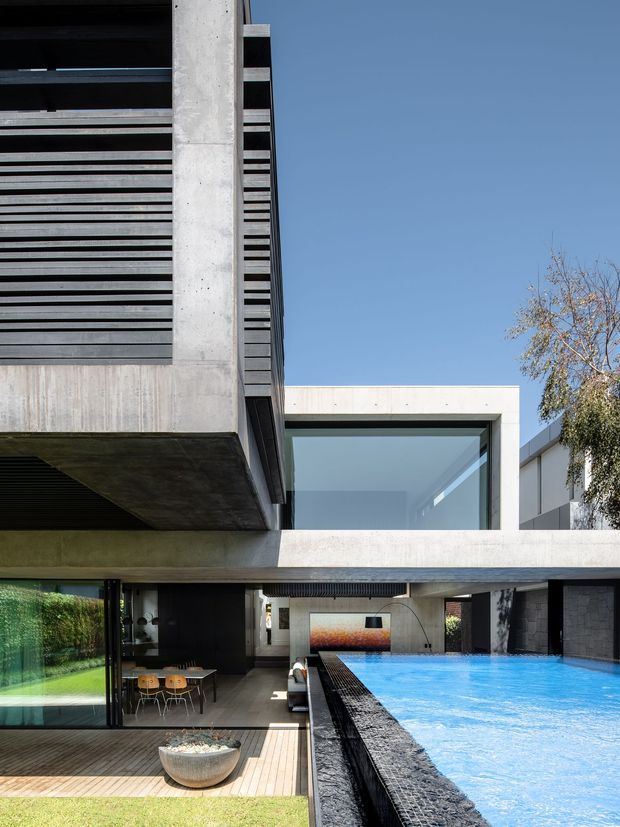 Contemporary multi-level concrete residence viewed from an outdoor pool area. The composition highlights strong horizontal lines, exposed concrete slabs and columns, dark louvred screening, expansive glazing, and a cantilevered upper volume. A vivid rectangular swimming pool occupies the foreground beside a timber deck, covered outdoor dining area, and landscaped garden.