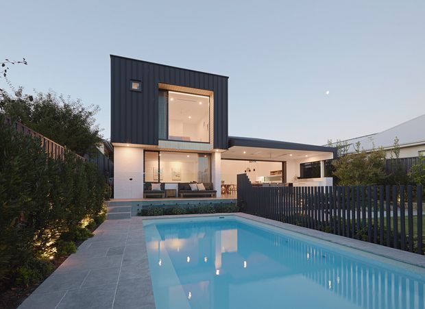 Contemporary two-storey residence viewed from the backyard at dusk, with dark vertical cladding, large glazed openings, a covered outdoor entertaining area, and a rectangular swimming pool surrounded by paved terraces and landscaped planting.