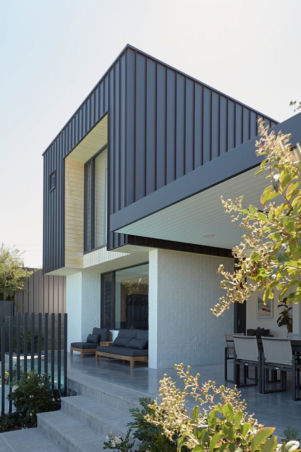 Exterior view of a contemporary multi-level residence with a dramatic steeply angled gabled roof form clad in dark vertical metal panels. The house features white painted brick walls, large glazed openings, a projecting covered terrace, and a pale linear soffit. Concrete steps lead to an outdoor living area furnished with a sofa and dining table, with landscaped planting in the foreground.