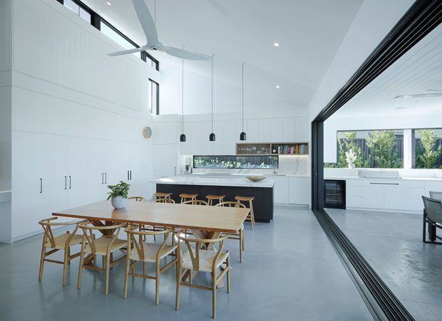 Bright open-plan contemporary kitchen and dining area with a long light-wood dining table and matching timber chairs in the foreground. White full-height cabinetry, a central island with bar seating, pale flooring, pendant lights, and high clerestory glazing create an airy space. A large black-framed glazed opening connects visually to an adjacent bright room with built-in cabinetry and a bench.
