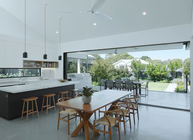 Bright, open-plan contemporary kitchen and dining area with a large dark island, white benchtops, pendant lighting, timber stools, and a long timber dining table. Full-width glazing opens the room visually to a leafy garden and outdoor entertaining area. The space has a clean minimalist fit-out, polished concrete-style flooring, and abundant natural light.