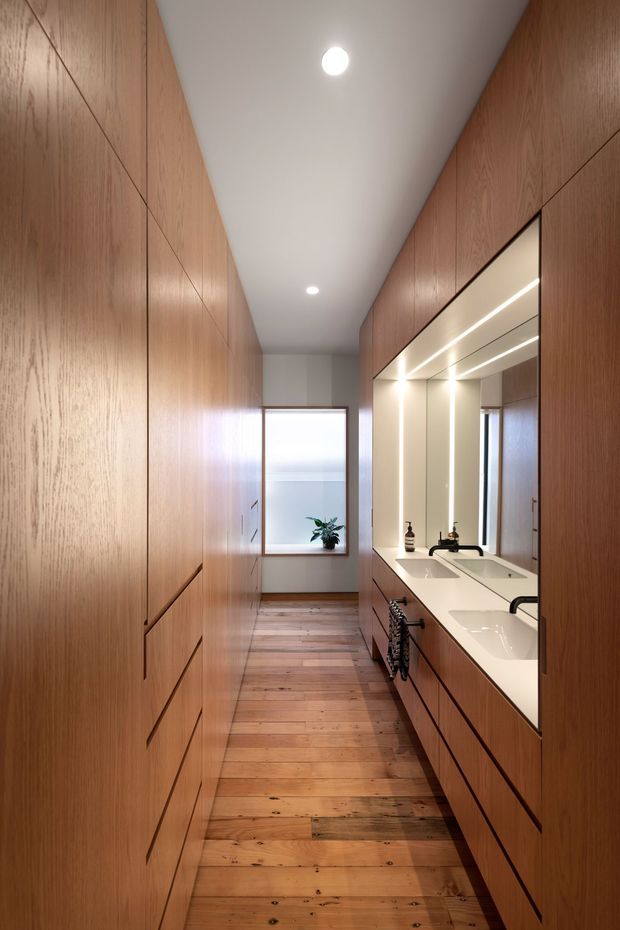A narrow, elongated bathroom corridor with extensive warm timber built-in cabinetry on both sides. The right side contains a long white double vanity with integrated rectangular basins, black wall-mounted taps, large mirrors, and concealed linear lighting. Timber plank flooring runs toward a bright window at the end, creating a warm and highly cohesive, minimalist interior.