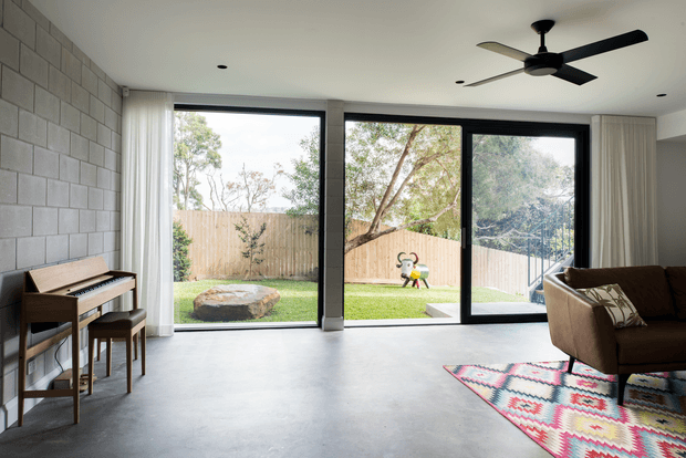 A bright contemporary living room with a polished concrete floor, exposed grey concrete block feature wall, full-height black-framed glazing and sliding doors opening to a fenced garden. The room includes a brown sofa, patterned rug, compact upright piano and ceiling fan, with abundant natural light and a simple, open layout.