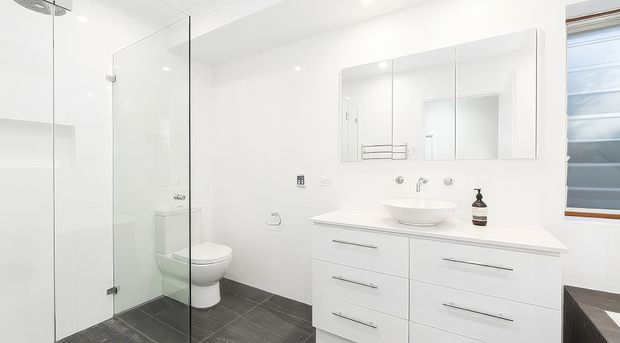Bright, contemporary white bathroom with a frameless glass shower enclosure, wall-hung toilet, floating white vanity with integrated drawers, round vessel basin, large mirrored cabinet, and dark grey tiled floor. A high horizontal window provides natural light.