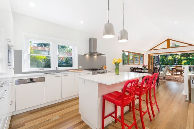 Bright open-plan kitchen with white cabinetry, stainless-steel appliances, a large island with a pale benchtop, and red bar stools. Timber flooring continues into an adjoining dining and living area, which is framed by extensive glazing and exposed timber roof structure. Pendant lights and garden views create an airy, contemporary family space.