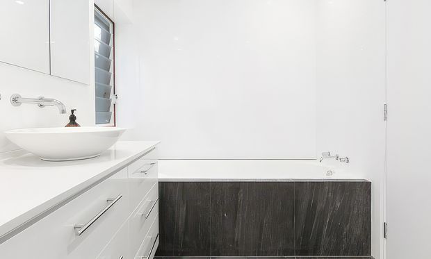 Bright, minimalist bathroom featuring a white vessel basin on a long white vanity with integrated drawers, a wall-mounted chrome tap, a louvred window, and a built-in bathtub enclosed by dark timber-look paneling. The room is predominantly white with clean, simple lines and strong contrast from the dark bath surround.