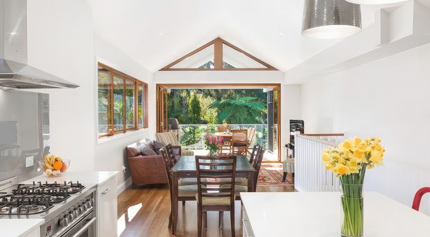 Bright open-plan kitchen and dining area with white cabinetry and benchtops, stainless-steel appliances, timber flooring, and a glazed dining extension opening to greenery. The dining space features a timber table and chairs, while a pitched timber-framed glazed roof element and large windows bring in abundant natural light.