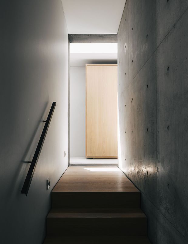 A narrow, minimalist stairway with dark timber treads, a simple black wall-mounted handrail, a smooth grey concrete feature wall, and white plaster walls. Natural light enters through a bright opening at the upper landing, highlighting a pale timber door or built-in panel and the contrasting raw concrete ceiling beam.
