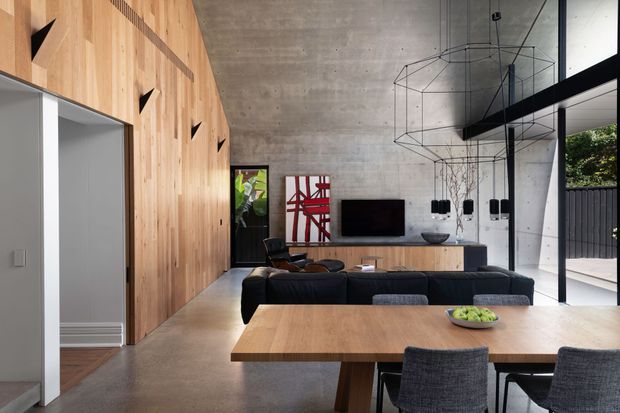A contemporary open-plan living and dining area with a dramatic double-height concrete ceiling and wall, extensive warm timber wall panelling, polished concrete flooring, and full-height glazing. A large timber dining table with upholstered chairs occupies the foreground, while a dark sectional sofa, low timber media console, television, sculptural pendant lighting, and indoor plants define the living zone. The space opens to an exterior terrace through large glass doors.