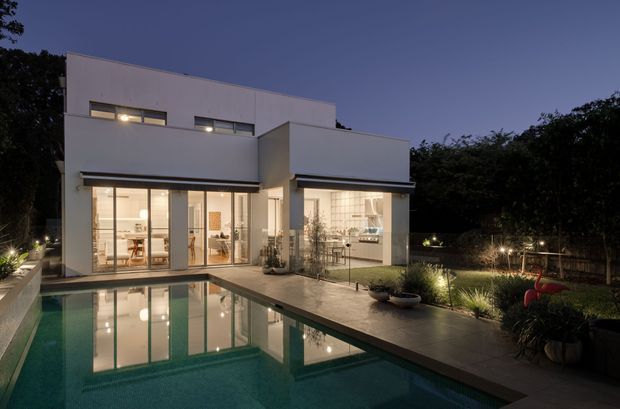 Twilight exterior view of a modern white two-storey residence with clean cubic volumes, extensive floor-to-ceiling glazing, and a flat roof. A rectangular swimming pool and paved terrace occupy the foreground, with landscaped planting and garden lighting surrounding the house. Interior living and dining areas are visible through the glazed openings.